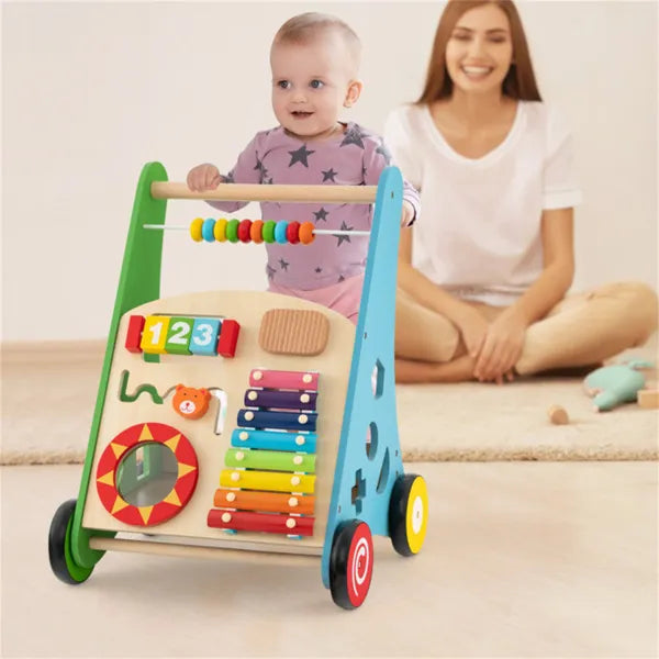 Child playing with a colorful wooden activity walker, with a woman sitting nearby.