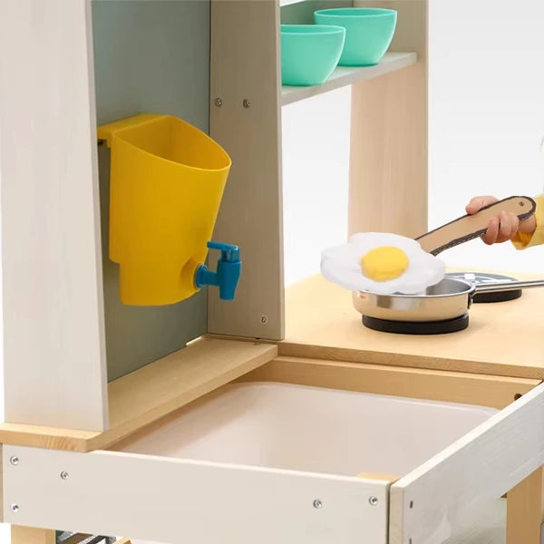 Children's play kitchen set with a yellow cup and blue spout, green bowls on shelves, and a hand holding a spatula.